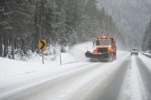 snow plow drives on snowy 2 lane highway through forest