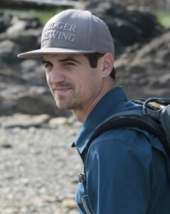 Mat Bell, a white man with dark hair and a short beard, looking away from the camera. He is wearing a backpack and ball cap and is on the edge of a river.