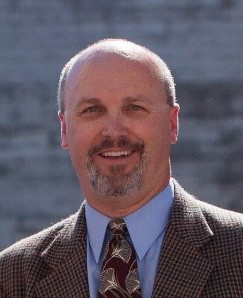 The portrait of a smiling White man, with short hair and a goatee, wearing a suit and tie.