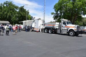 A dump truck pulling an enclosed trailer with metal towers erected at either end. People are gathered to view the inside of the trailer.