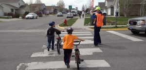 Two middlesschool students push bicycles across a pedestrian crossing while a crossing guard monitors traffic
