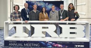 Seven adults in their 20s and 30s stand behind a sign that says TRB annual meeting January 7-11 2024