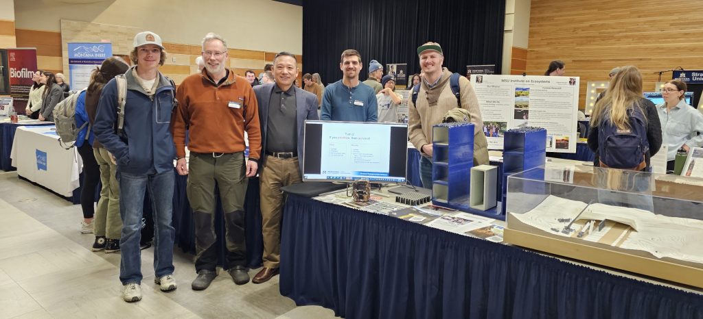 Marcel Huijser Kelvin Wang and Mat Bell pose for a photo with two MSU undergraduate students. They stand next to a table with a wildlife crossing model fiber reinforced polymer blocks and a PowerPoint display.