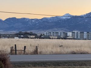 Urban fringe view with open field and grazing deer in the foreground, urban housing and commercial in the mid-ground and mountains with sunrise in the foreground.